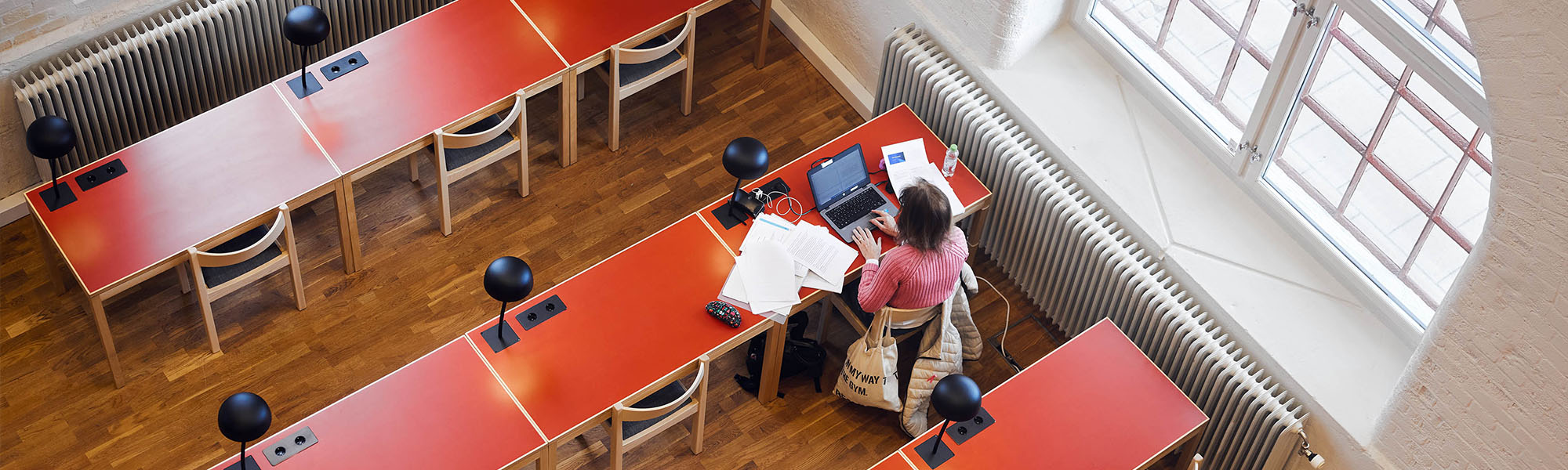 A lone student studying with a laptop and lots of documents spread out on a red table. The table is next to a large window. The picture is taken from above.